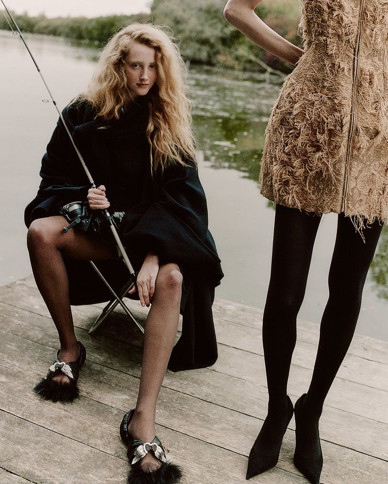 Two women pose on a wooden dock over a lake. One woman sits with a fishing pole. Editorial fashion shot with outdoor scenic background.