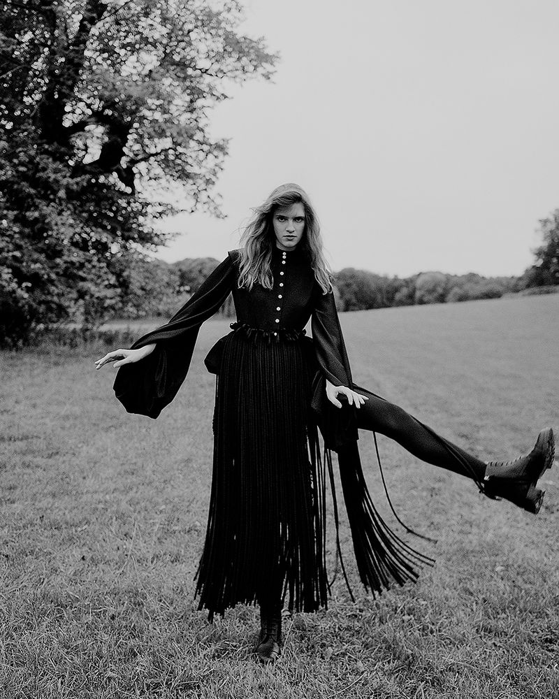 Monochrome fashion editorial featuring a woman in a dark fringed dress posing on an outdoor grassy field with trees