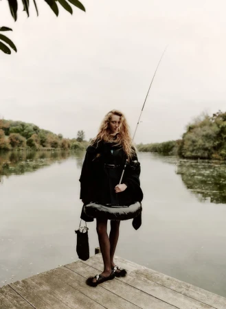 A woman in an oversized coat standing on a wooden pier next to a lake holding a fishing rod on a cloudy day in a rural setting.