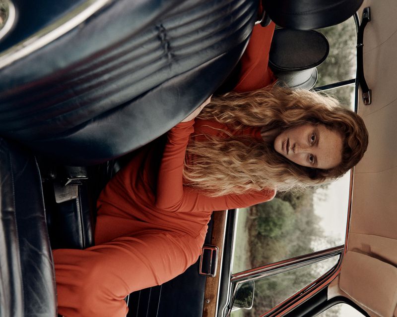 An adult woman with long curly hair sits in the back of a vintage car wearing a red dress, creating a retro and elegant fashion statement.