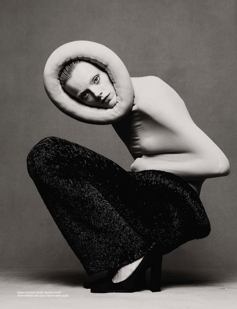 Fashion shot of a woman crouching, wearing black trousers and a beige neckpiece, in a studio with a gray background and neutral lighting.
