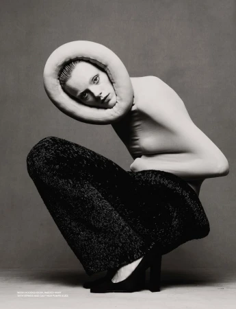 Fashion shot of a woman crouching, wearing black trousers and a beige neckpiece, in a studio with a gray background and neutral lighting.