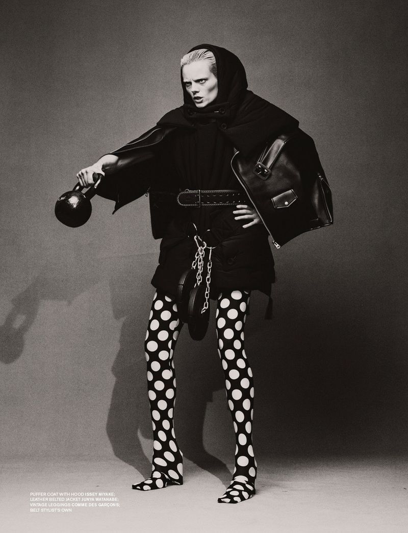 Monochrome fashion shot of a model holding a kettlebell. She wears a headscarf, jacket, polka dot tights, and a belt.