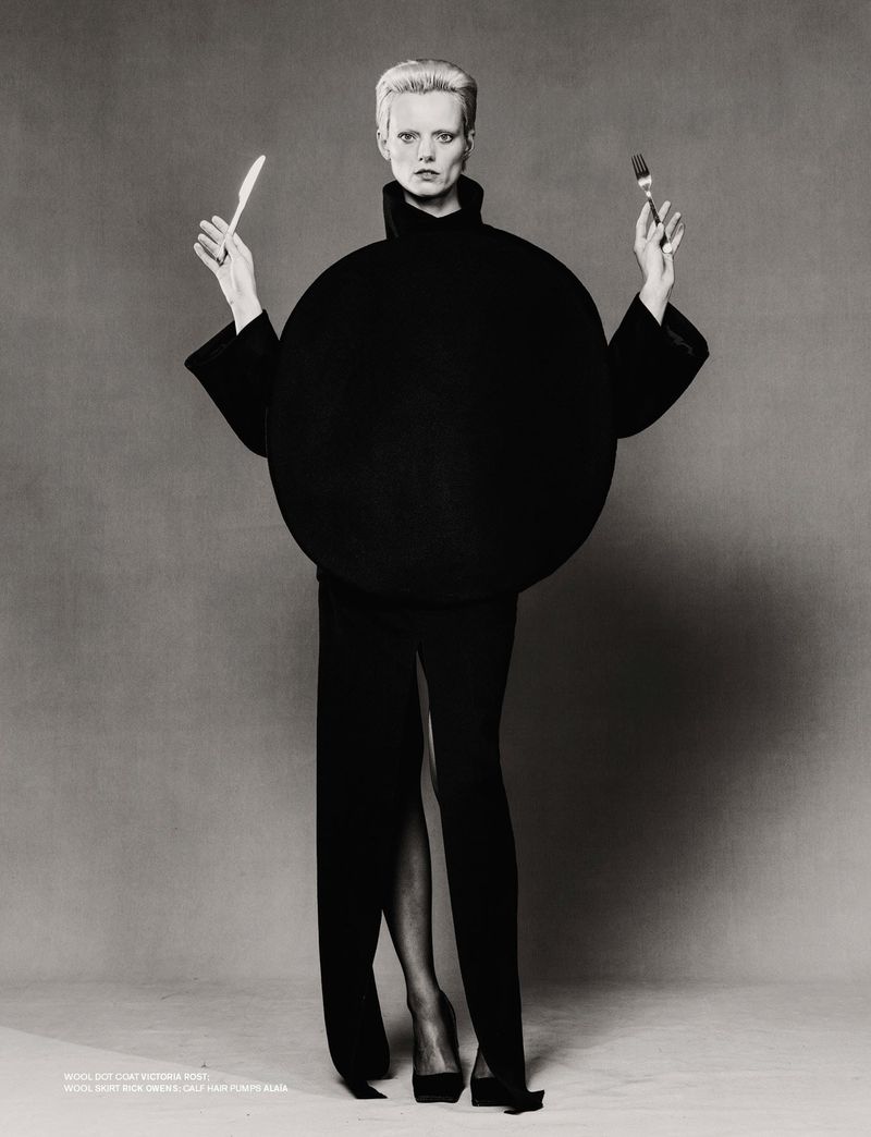 A black-and-white fashion shot of a woman in a black dress with a large round shape, holding a knife and fork, standing in a studio.