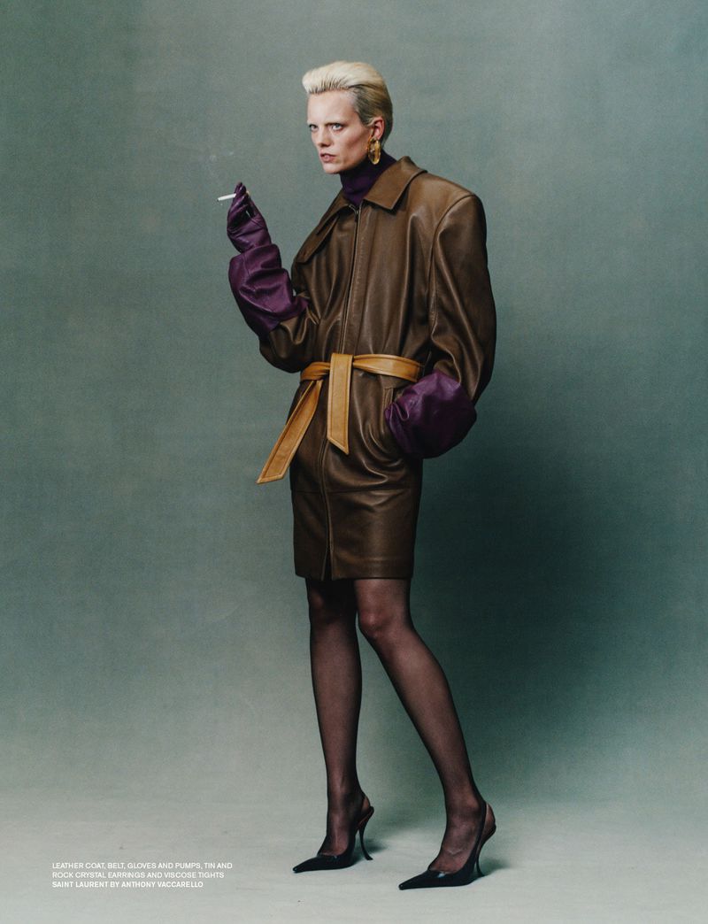 A fair-skinned model poses in a brown leather dress with oversized sleeves, a belt, black hosiery, and high heels, smoking a cigarette.