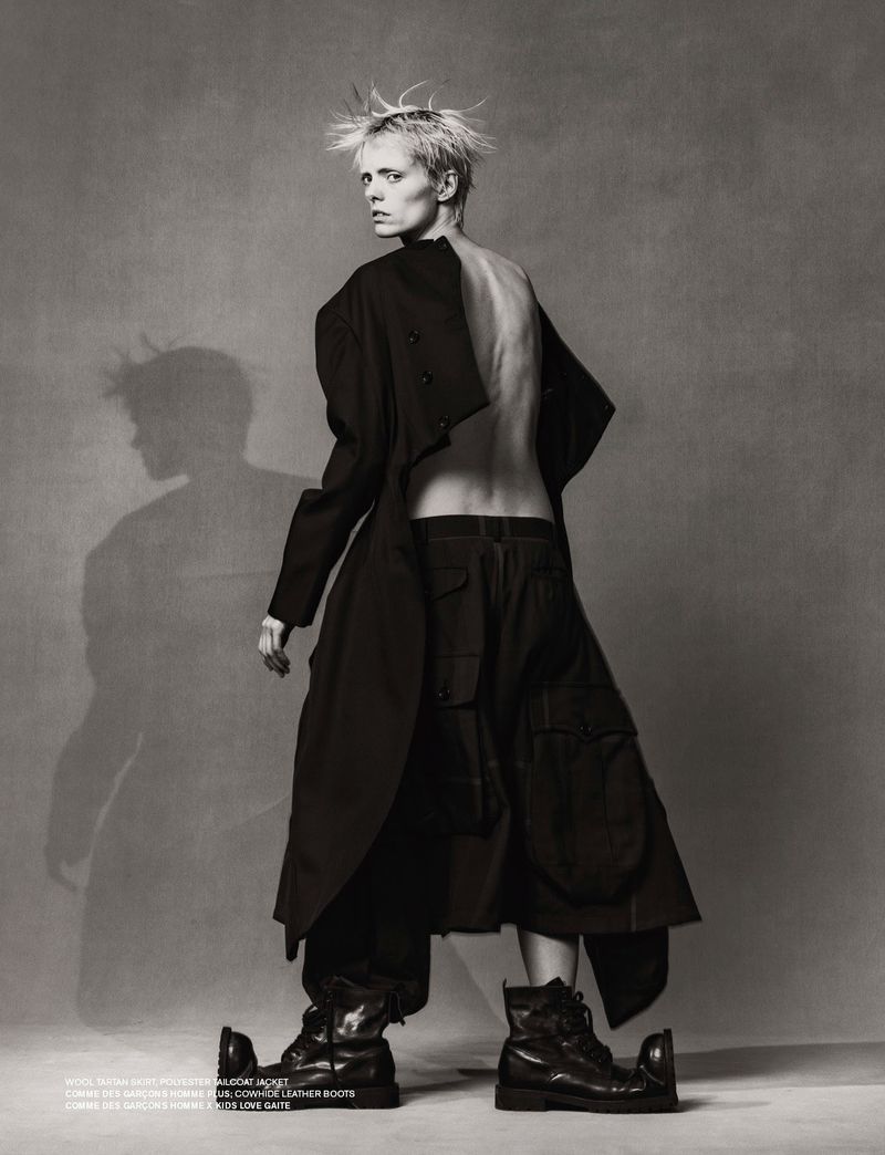 Fashion editorial shot of a thin adult woman in an oversized black jacket and boots, standing indoors against a grey background.