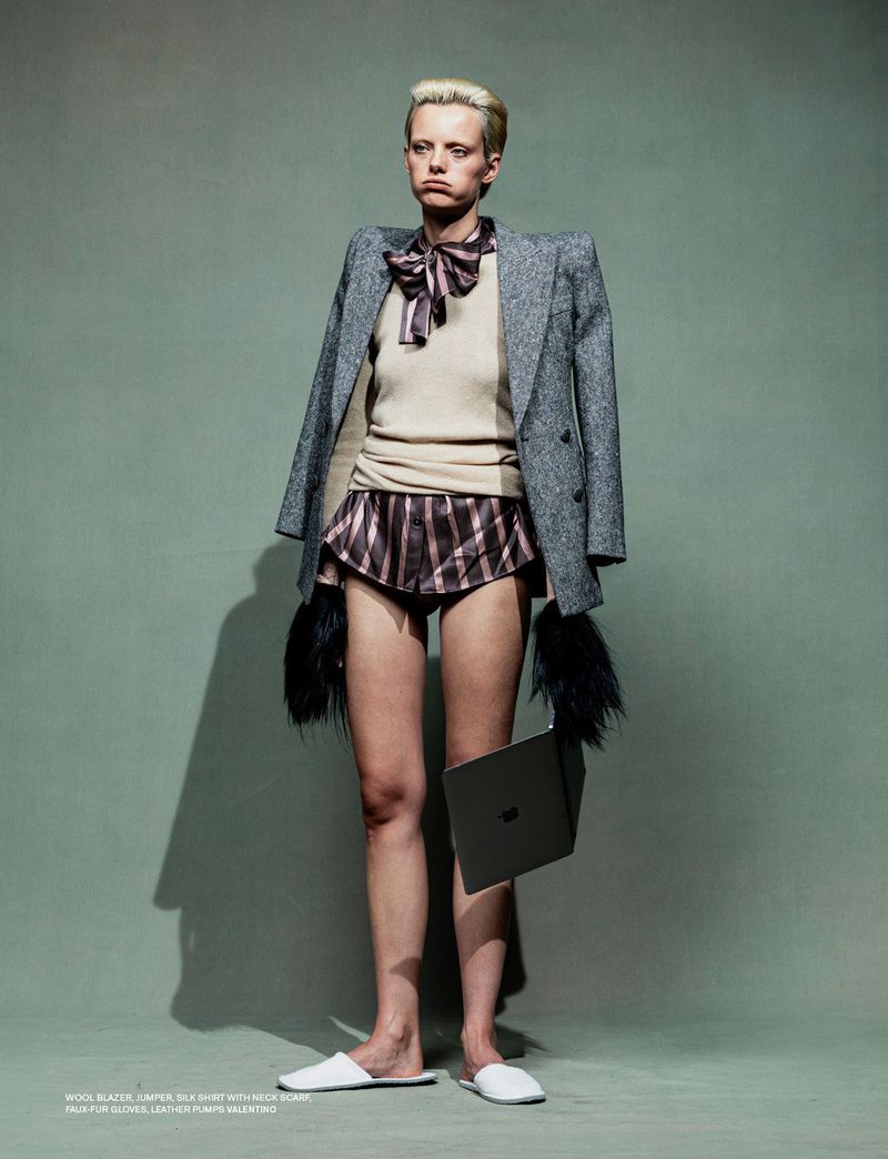Fashion portrait of a woman wearing blazer, sweater, striped shorts, and slippers, holding a laptop in a studio setting.