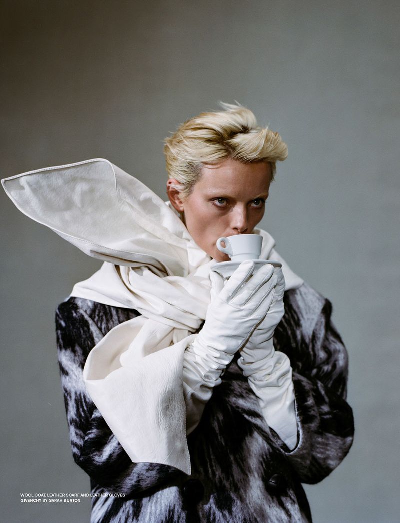 Close-up portrait of a person drinking from a teacup, wearing white gloves, a scarf, and a black and white patterned wool coat indoors.