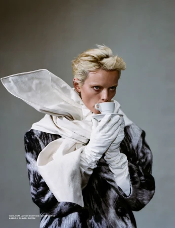 Close-up portrait of a person drinking from a teacup, wearing white gloves, a scarf, and a black and white patterned wool coat indoors.