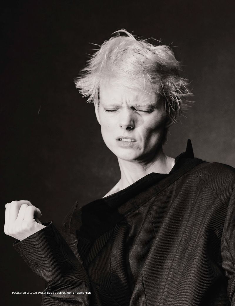 Monochrome portrait of a fashion model with short blonde hair wearing a dark jacket, eyes closed, and face scrunched in concentration.