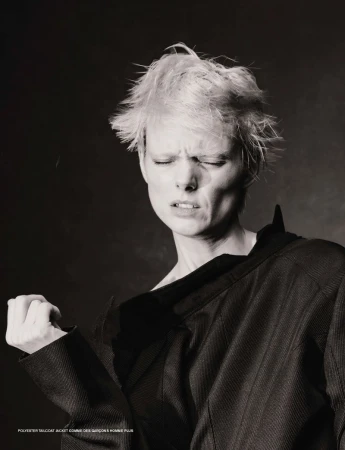 Monochrome portrait of a fashion model with short blonde hair wearing a dark jacket, eyes closed, and face scrunched in concentration.