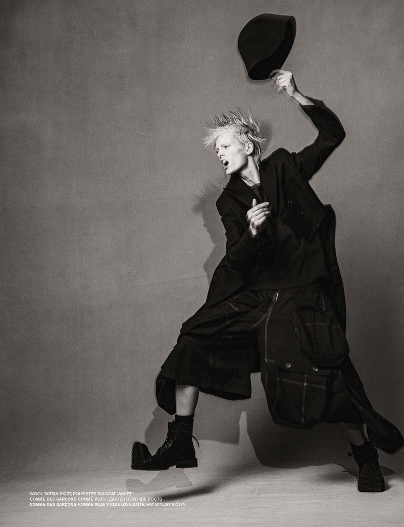 Monochrome fashion shot of a fair-skinned model in a dark, oversized jacket and culottes, tossing a hat, against a textured backdrop.