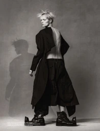 Model with spiky blonde hair wearing an open-back black coat and dark boots posing against a neutral textured studio background.