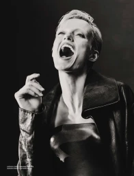 Black and white portrait of a laughing woman wearing a black leather corset top and leather jacket against a dark studio backdrop.