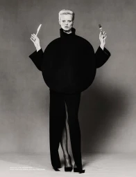 Model with blonde hair holding a knife and fork wearing a structured round black garment against a grey studio backdrop.