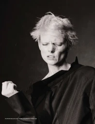 Model Maaike Klaasen in an oversized black jacket with eyes closed and tousled bleached hair against a dark studio background.