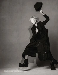 Model Maaike Klaasen mid-air in a dynamic jump holding a black hat, styled in an oversized dark coat and boots against a studio backdrop.