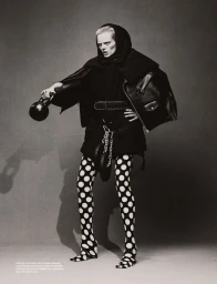 Model posing in a structured black hooded jacket and polka dot tights while holding a kettlebell against a grey studio backdrop.