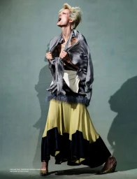 Model with short blonde hair yelling while clutching a draped grey fabric top and wearing a yellow and black skirt against a muted background