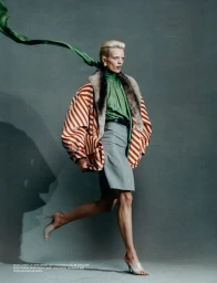Maaike Klaasen mid-stride wearing an orange striped jacket with fur collar, green silk top with flowing sash, and grey pencil skirt.