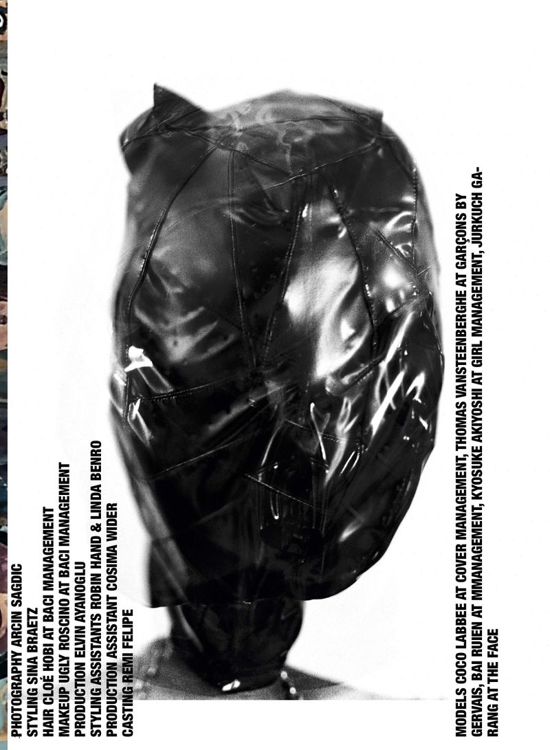 Black and white studio fashion shot of a figure fully covered with a shiny black plastic hood, photographed against a stark white background.
