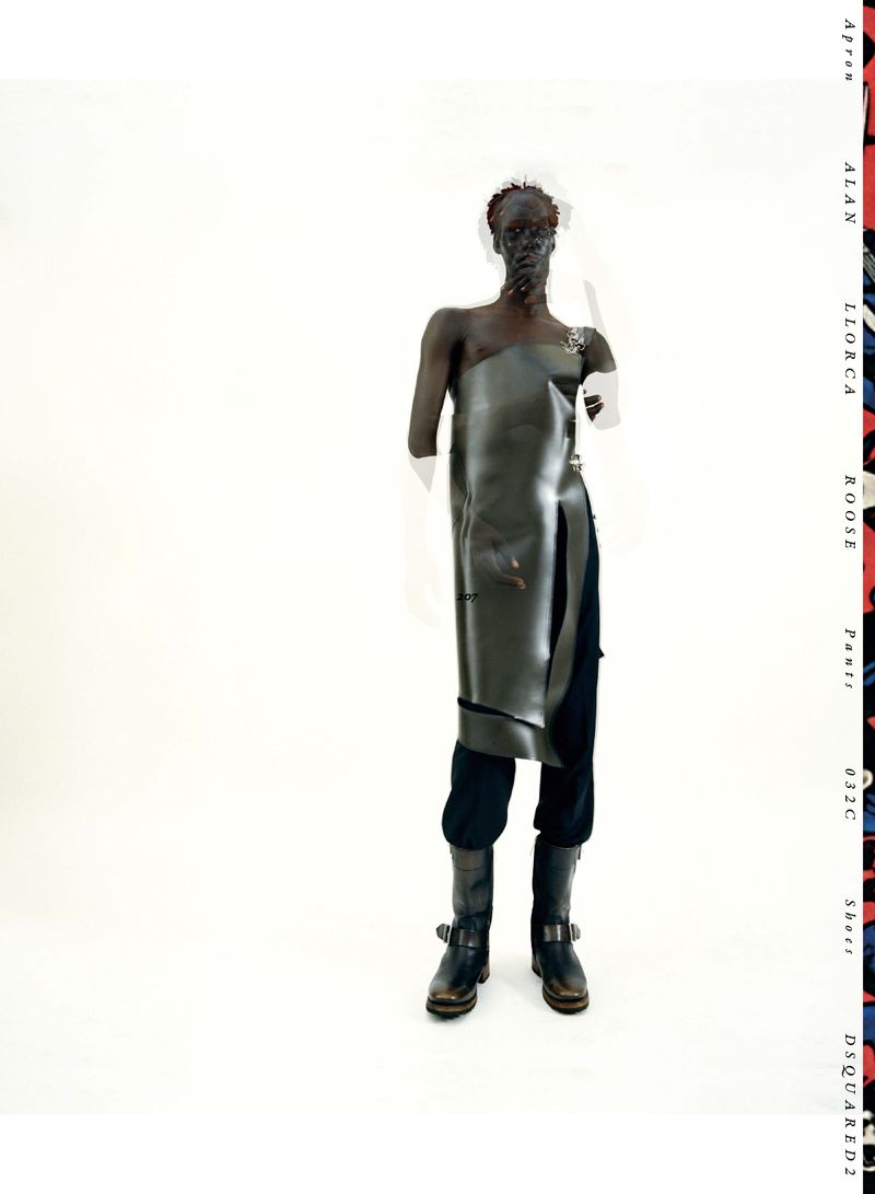 Editorial fashion shot of a slender adult with dark skin in a vinyl apron, black pants, and boots against a white backdrop with column text.
