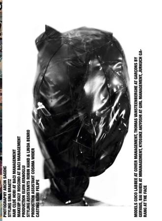 Model wearing a glossy black full-head mask covering the face, photographed against a stark white seamless backdrop.