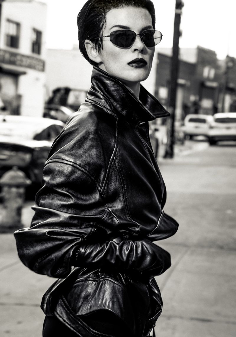 A striking black and white portrait of a woman in a leather jacket and sunglasses standing on a sidewalk in an urban setting.