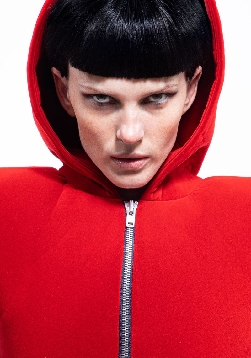 Close-up portrait of a person with short black hair and pale skin, wearing a bright red hooded jacket with a silver zipper on a white background.