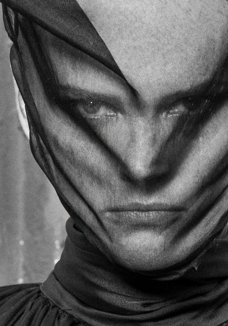 Monochrome portrait of adult woman covered in sheer black fabric, gazing at camera with a serious expression, against an out-of-focus background.