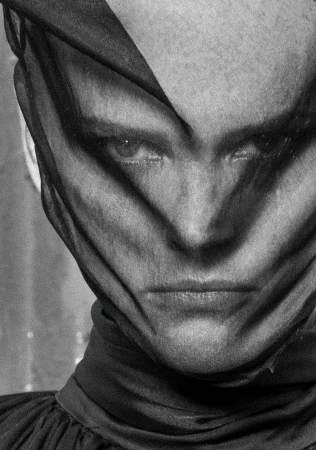 Monochrome portrait of adult woman covered in sheer black fabric, gazing at camera with a serious expression, against an out-of-focus background.
