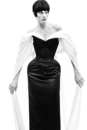Elegant woman in a black satin dress with white chiffon sleeves, standing against a bright white background in a fashion forward pose.