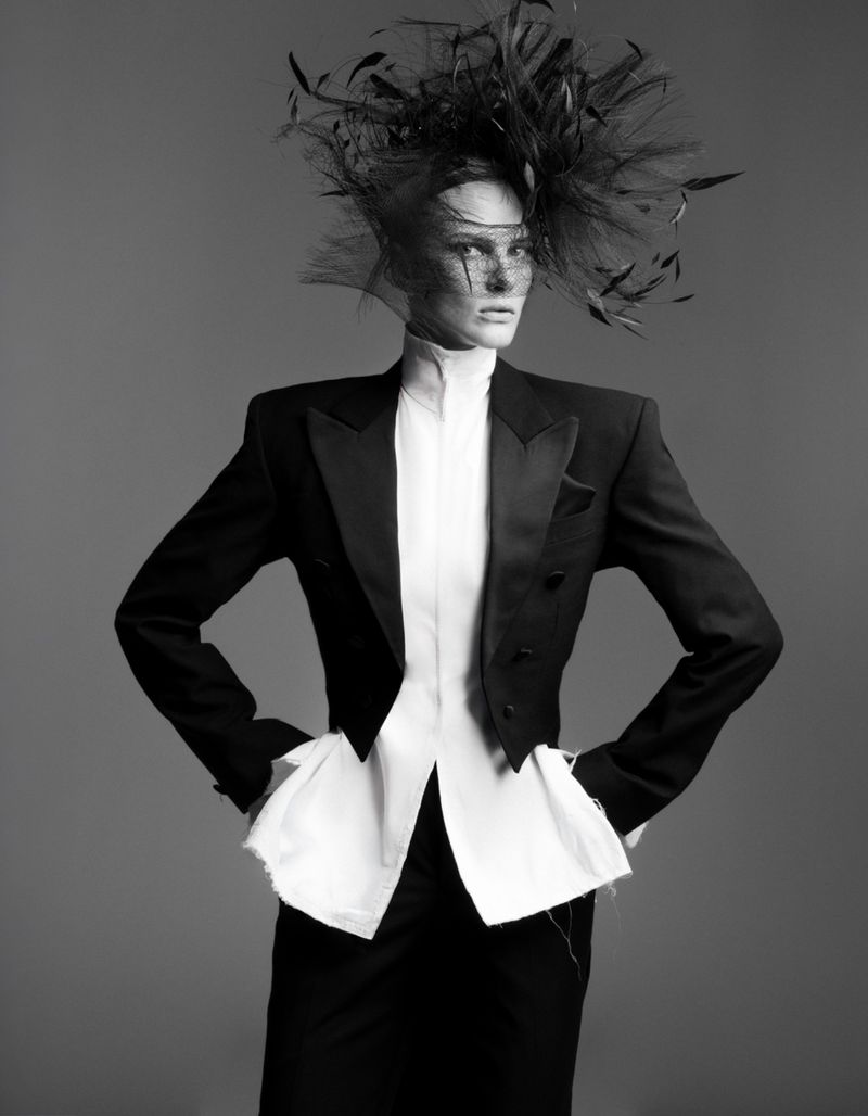 Elegant model in black tuxedo jacket and feathered headpiece poses in a studio portrait. Striking monochrome fashion shot with dramatic flair.