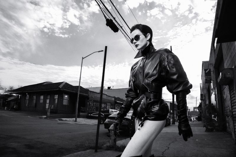 Black and white fashion portrait of a woman in a leather jacket and sunglasses walking on an urban street with buildings and utility poles.