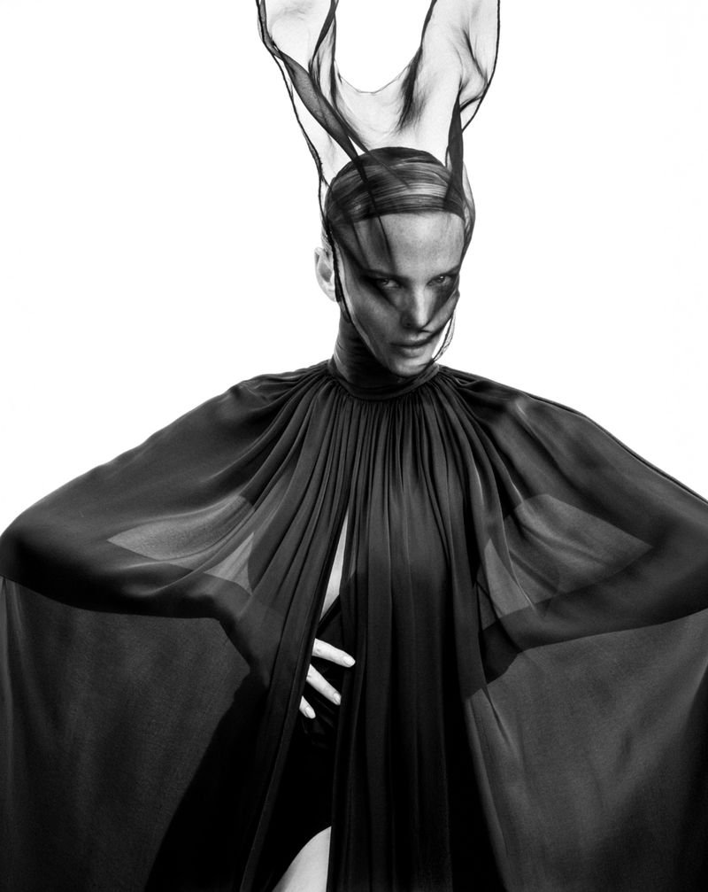 Striking black and white fashion portrait of a model in a dark sheer gown and head covering against a stark white backdrop.