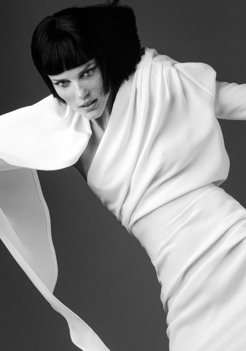 Elegant studio portrait of a female model with dark hair, wearing a flowing white dress, against a dark grey background, emphasizing fashion.