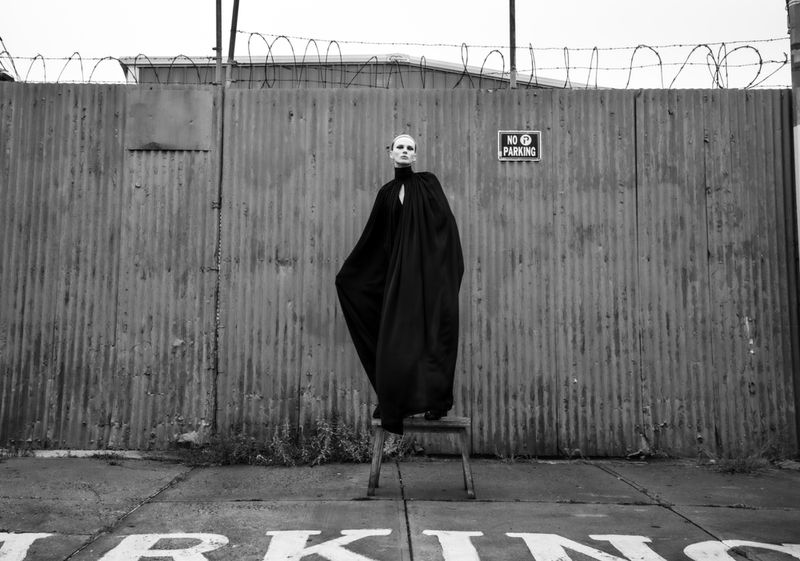 Monochrome fashion shot of a slender woman in a black cape standing on a stool in front of a corrugated metal wall with barbed wire.