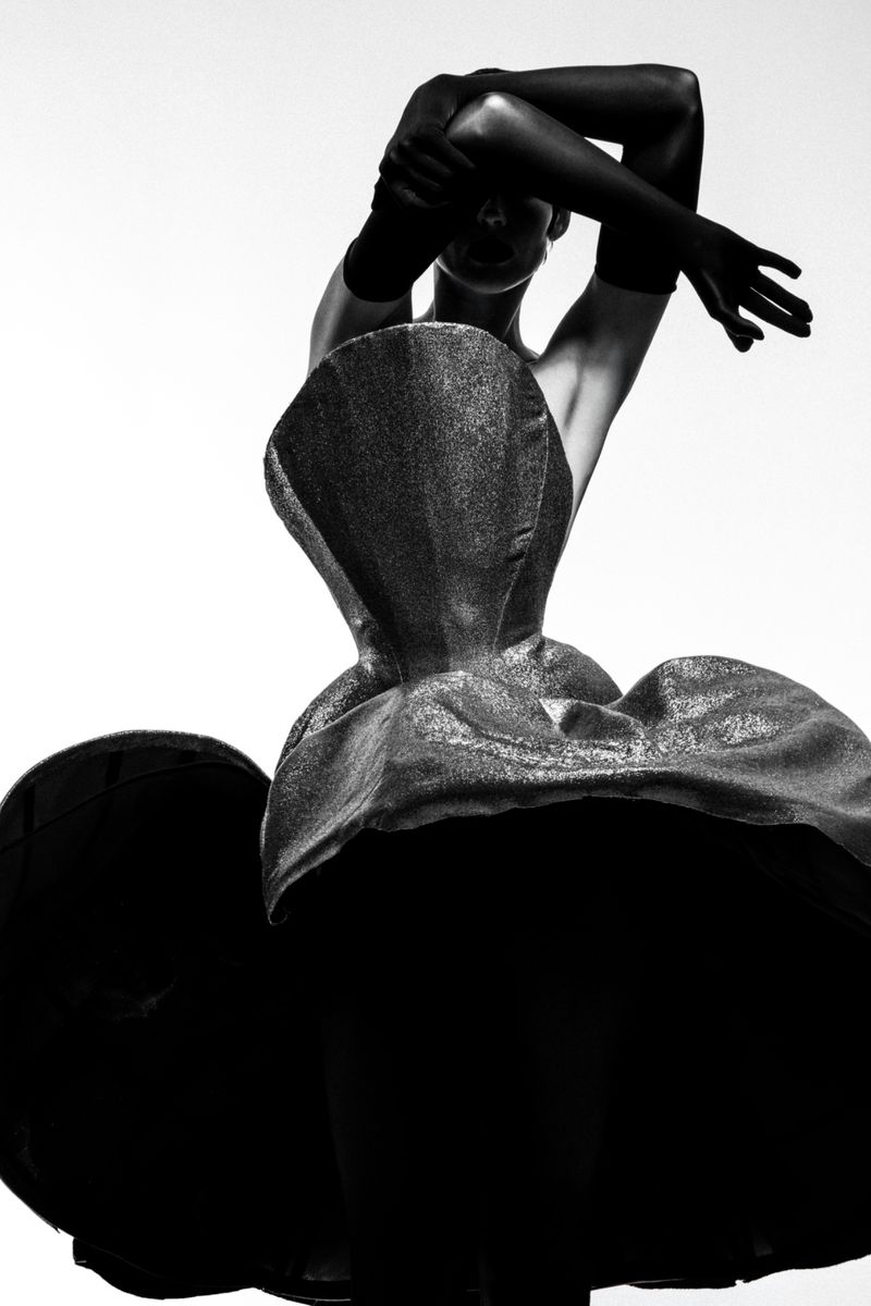 Artistic black and white fashion portrait of a person in a sculpted dress and gloves, posing in a studio with a bright background.
