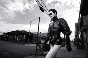 Anne Vyalitsyna walking down a city street wearing an oversized black leather jacket, gloves, and sunglasses in black and white.