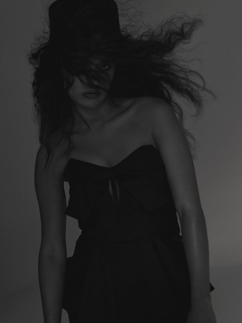 Portrait of Asha Banks wearing a black strapless top and top hat with hair blowing across her face against a dark studio background
