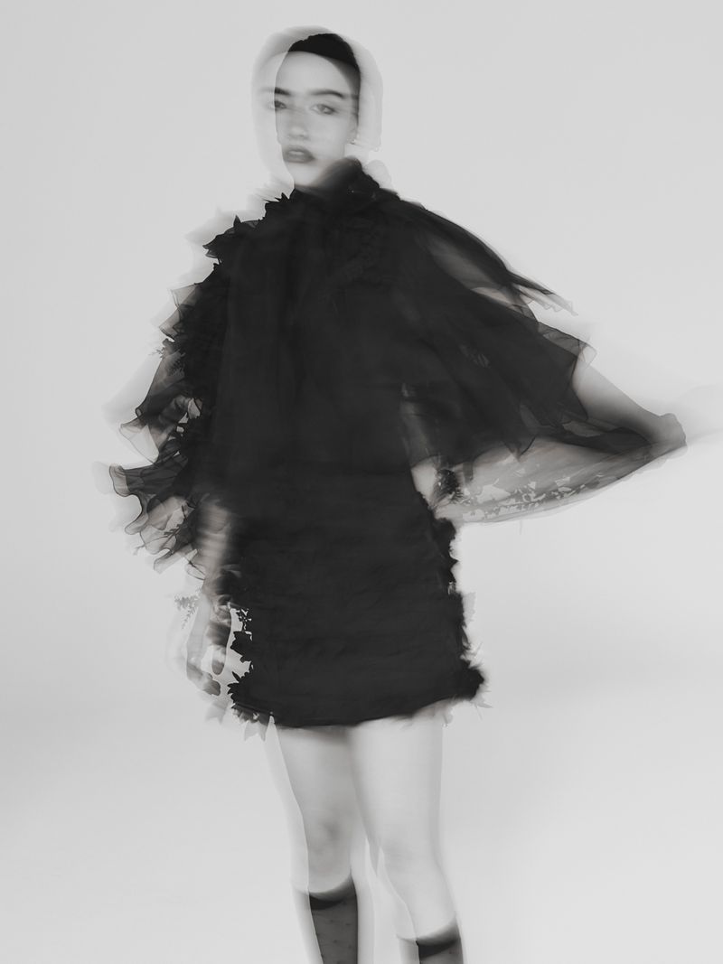 Fashion shot of a blurry young woman in a black ruffled dress and sheer headscarf against a white backdrop, exhibiting a sense of motion.