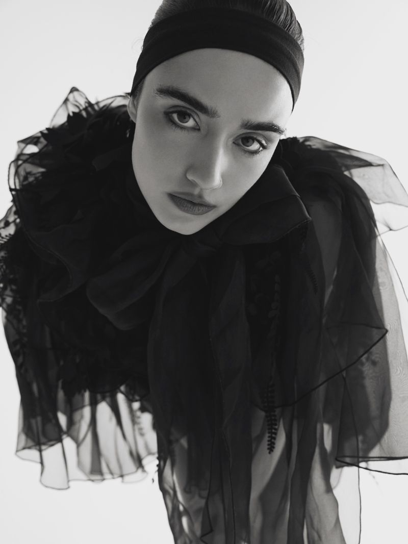 Striking black-and-white portrait of a young adult woman with a headband, wearing a frilled sheer black top, set against a clean white backdrop.
