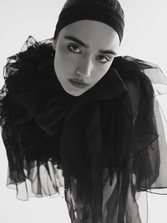 Striking black-and-white portrait of a young adult woman with a headband, wearing a frilled sheer black top, set against a clean white backdrop.