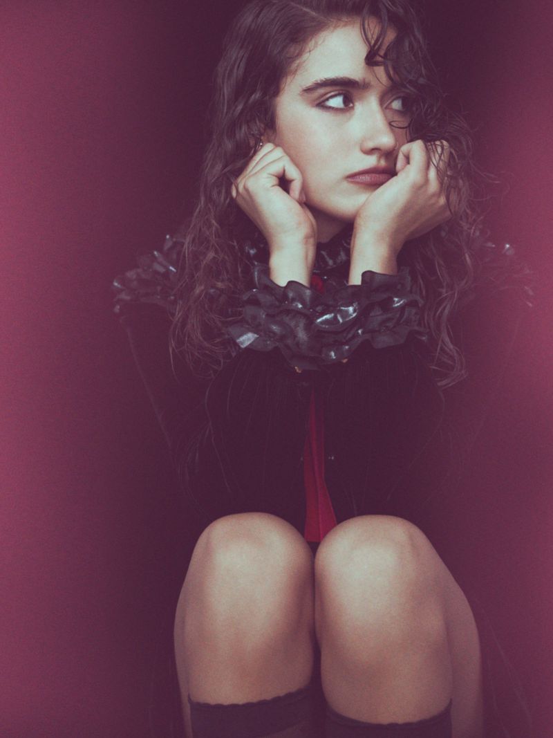 Young woman with curly brown hair sits with knees up and hands at her face in a portrait shot against a red background.
