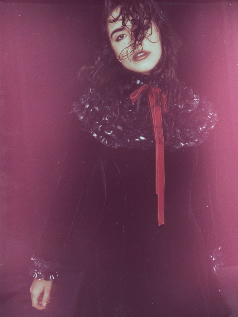 A young woman with dark hair and a dramatic ruffled collar poses in a dark velvet dress against a deep pink studio backdrop in a fashion editorial.