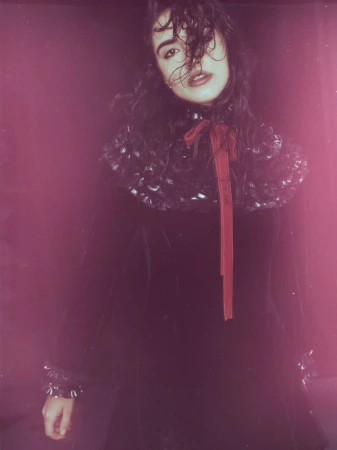 A young woman with dark hair and a dramatic ruffled collar poses in a dark velvet dress against a deep pink studio backdrop in a fashion editorial.