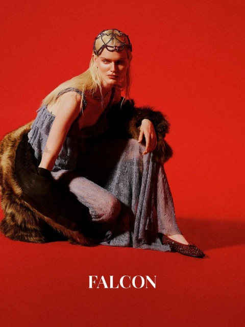 Fashion photo of a person with long blonde hair wearing a dress, fur stole, and beaded headpiece against a red background, studio shot.