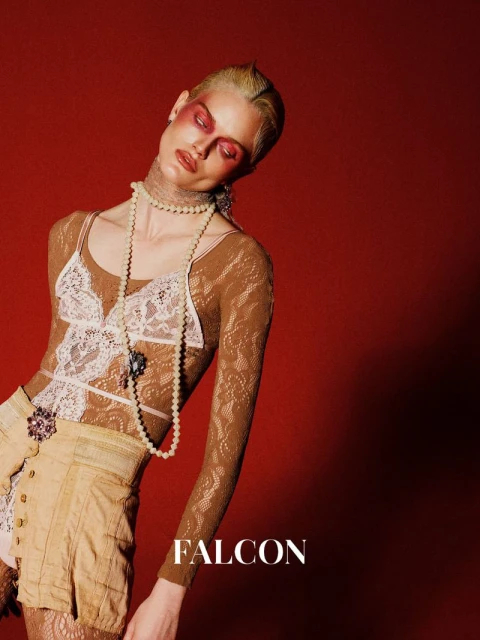 A woman with avant-garde makeup and a pearl necklace poses against a red backdrop wearing a mesh top and khaki shorts.