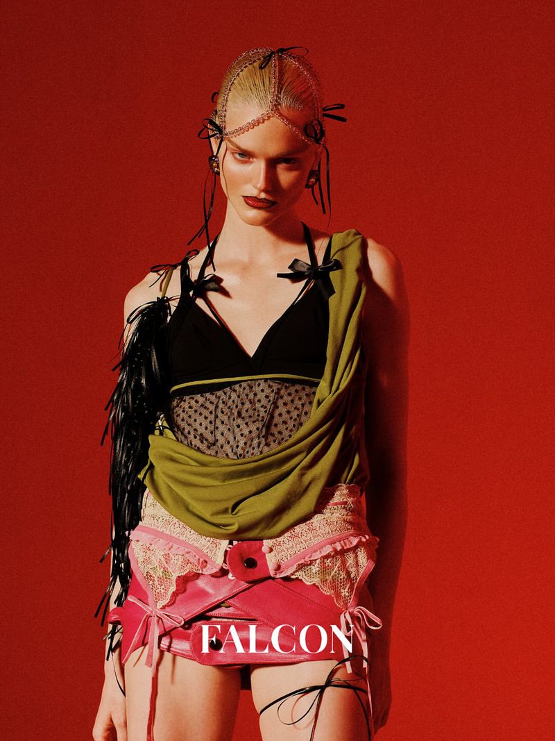 A person is posing in a studio with a red background, wearing a headpiece, a black top with straps, and a pink and beige skirt.