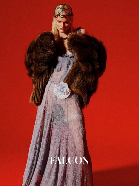 A blonde woman poses in a long dress and fur stole against a red background in a fashion-style portrait, wearing an embellished headpiece.
